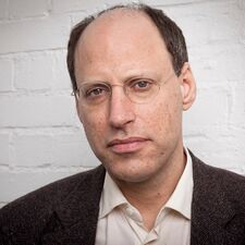 Portrait of Darian Leader: a man with glasses and brown hair in front of a white brick wall