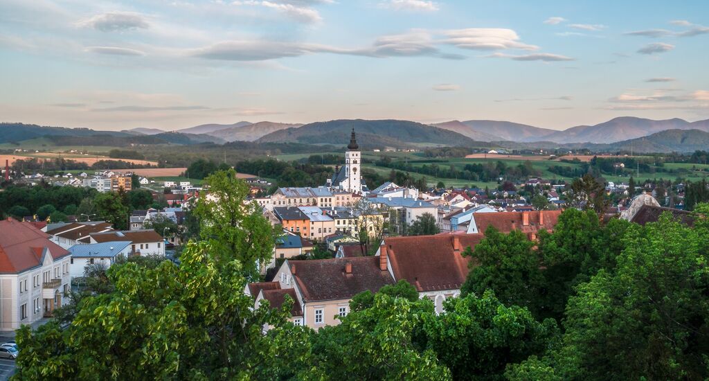 Eine Stadt mit prägnantem Kirchturm, Bäumen im Vordergrund, Bergen im Hintergrund